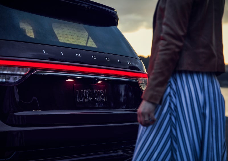A person is shown near the rear of a 2024 Lincoln Aviator® SUV as the Lincoln Embrace illuminates the rear lights | Buss Lincoln in McHenry IL